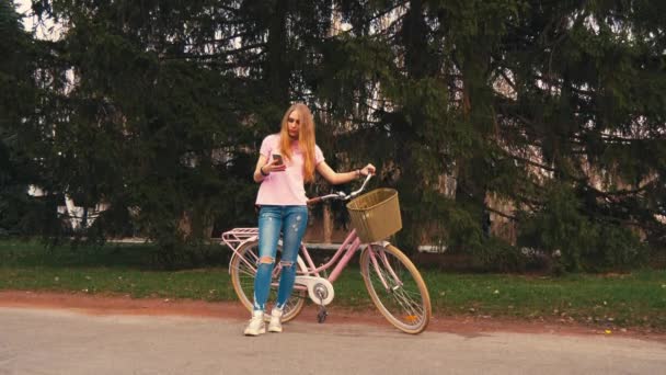 Ginger ruda dziewczyna siedzi na rower i SMS-y na smartfonie w lato park. Na sobie różową koszulę i spodnie — Wideo stockowe