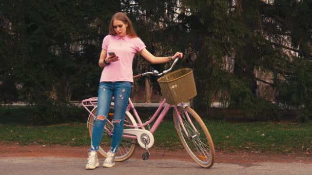 Імбирна руда дівчина сидить на велосипеді і надсилає смс на смартфон у літньому парку. Носіння рожевої сорочки та джинсів — стокове відео