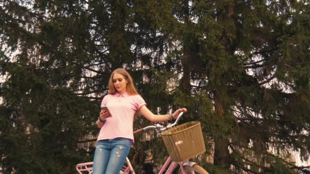 Ginger redhead girl sitting on the bike and texting on smartphone in summer park. Wearing pink shirt and jeans — Stock Video