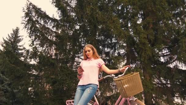Chica pelirroja pelirroja sentada en la bicicleta y mensajes de texto en el teléfono inteligente en el parque de verano. Usar camisa rosa y jeans — Vídeos de Stock