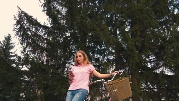 红头发的女孩坐在自行车上, 在夏季公园的智能手机上发短信。穿着粉色衬衫和牛仔裤 — 图库视频影像