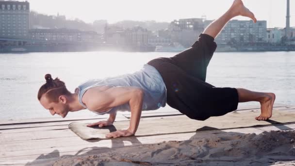 Młody człowiek kaukaski relaksujący praktykując ćwiczenia fitness joga na plaży, w pobliżu spokojna rzeka z miastem w tle — Wideo stockowe