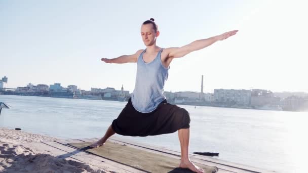 Młody człowiek kaukaski relaksujący praktykując ćwiczenia fitness joga na plaży, w pobliżu spokojna rzeka z miastem w tle — Wideo stockowe
