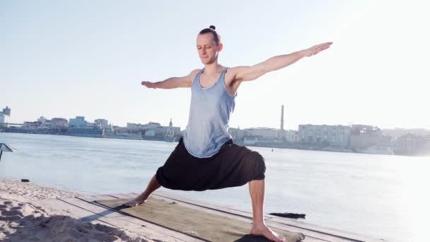 Kaukasiska ung avkopplande genom att praktisera yoga fitness motion på stranden nära lugn flod med staden på bakgrunden — Stockvideo