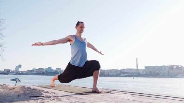 Hombre caucásico joven que se relaja practicando ejercicios de yoga en la playa cerca del río tranquilo con la ciudad al fondo — Vídeos de Stock