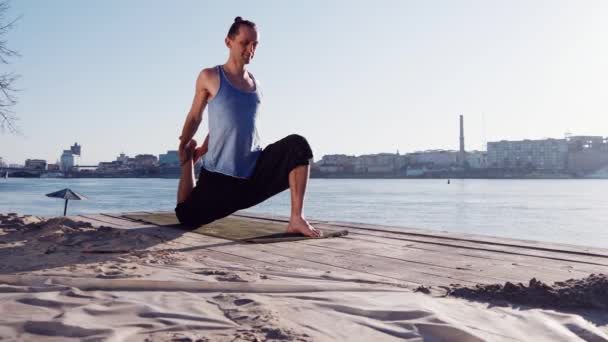 Hombre caucásico joven que se relaja practicando ejercicios de yoga en la playa cerca del río tranquilo con la ciudad al fondo — Vídeos de Stock