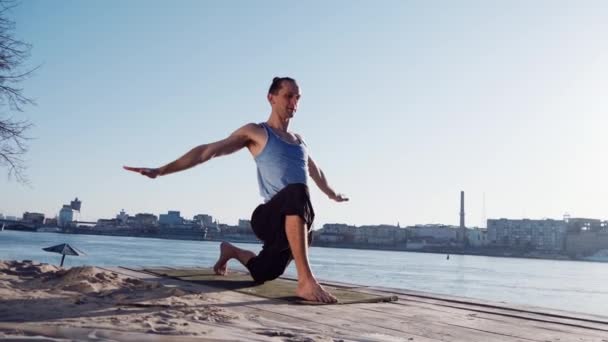 Kaukasische jongeman ontspannen door het beoefenen van yoga fitness oefening op het strand in de buurt van kalme rivier met stad op de achtergrond — Stockvideo