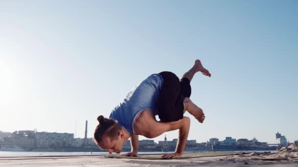 Jovem caucasiano relaxante praticando ioga exercício de fitness na praia perto de rio calmo com a cidade no fundo — Vídeo de Stock