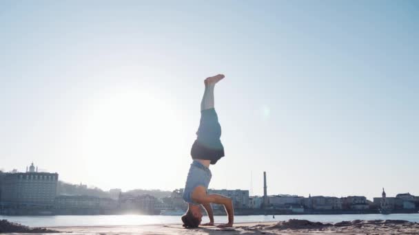 Kaukasiska ung avkopplande genom att praktisera yoga fitness motion på stranden nära lugn flod med staden på bakgrunden — Stockvideo