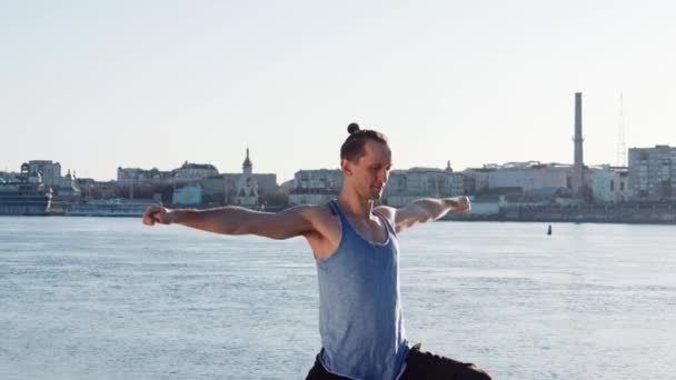 Kaukasische jongeman ontspannen door het beoefenen van yoga fitness oefening op het strand in de buurt van kalme rivier met stad op de achtergrond — Stockvideo