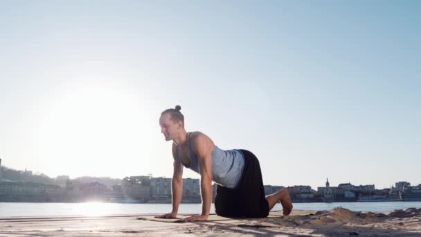 Hombre caucásico joven que se relaja practicando ejercicios de yoga en la playa cerca del río tranquilo con la ciudad al fondo — Vídeos de Stock