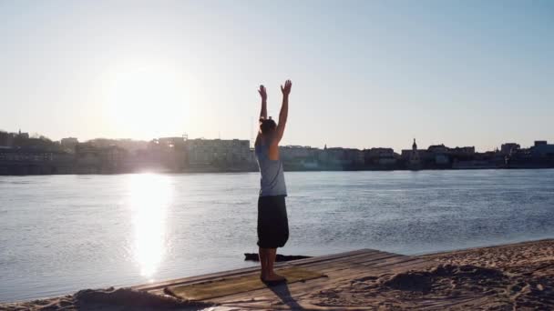 Молодой кавказский мужчина расслабляется, практикуя йогу фитнес-упражнения на пляже возле спокойной реки с городом на заднем плане — стоковое видео