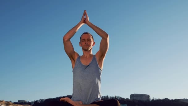 Молодой кавказский мужчина расслабляется, практикуя йогу фитнес-упражнения на пляже возле спокойной реки с городом на заднем плане — стоковое видео