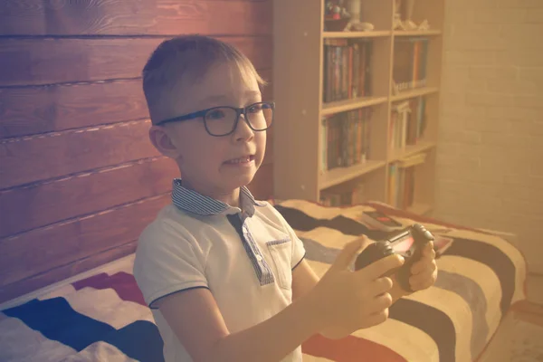 Caucásico chico en gafas sentado en su habitación en la cama con un joystick en las manos . —  Fotos de Stock