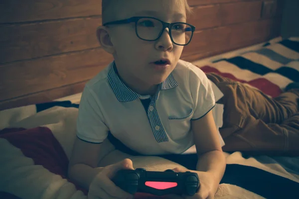 Niño caucásico en gafas acostado en la cama de su habitación con un joystick en las manos. Juega videojuegos en una consola —  Fotos de Stock