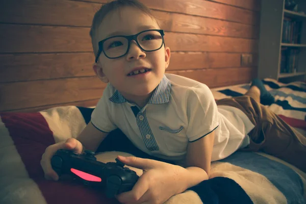 Biały chłopiec w okularach leżący na łóżku w swoim pokoju z joystickiem w rękach. Gra w gry wideo na konsoli. — Zdjęcie stockowe