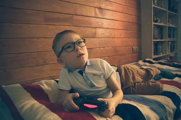 Niño caucásico en gafas acostado en la cama de su habitación con un joystick en las manos. Juega videojuegos en una consola —  Fotos de Stock