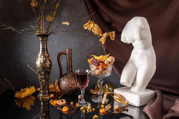 Still life with a plaster bust of Venus on a background of drapery, branches with autumn leaves in a vase, dried fruits and red wine — Stock Photo, Image