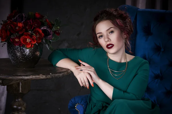 Young beautiful red-haired caucasian woman with professional makeup in green dress posing on blue sofa — Stock Photo, Image