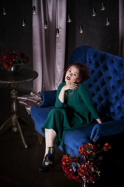 Молодая красивая рыжая белая женщина с профессиональным макияжем в зеленом платье позирует на синем диване — стоковое фото