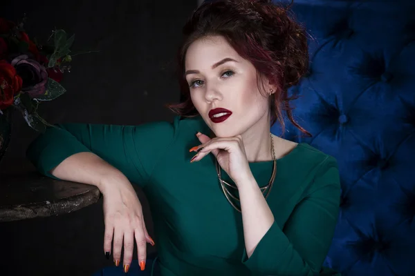 Young beautiful red-haired caucasian woman with professional makeup in green dress posing on blue sofa — Stock Photo, Image