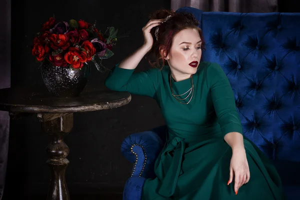 Young beautiful red-haired caucasian woman with professional makeup in green dress posing on blue sofa — Stock Photo, Image