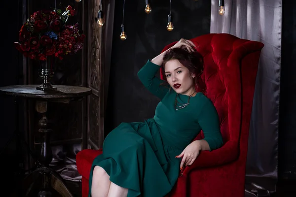 Giovane bella donna caucasica dai capelli rossi con trucco professionale in abito verde in posa sul divano rosso — Foto Stock