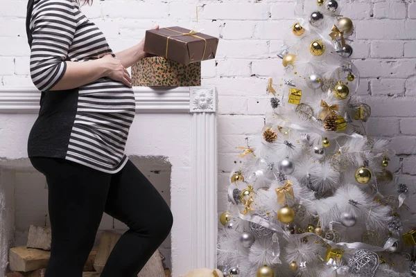Mladá těhotná žena s krabičky pózuje na pozadí vánoční stromeček — Stock fotografie