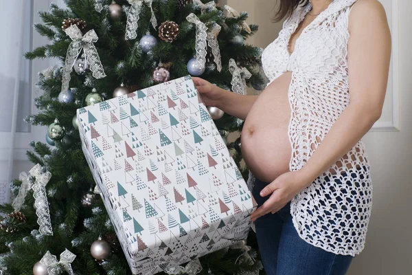 Young pregnant woman with gift box posing on the background of Christmas tree — Stock Photo, Image