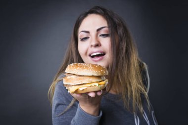 Happy beautiful young plus size model posing with hamburger on a gray studio background, xxl woman eating burger clipart