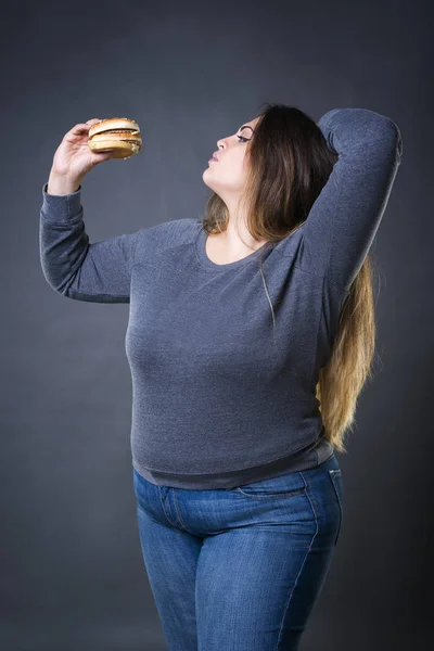 Hermosa joven modelo de tamaño más posando con hamburguesa sobre un fondo de estudio gris, xxl mujer comiendo hamburguesa — Foto de Stock