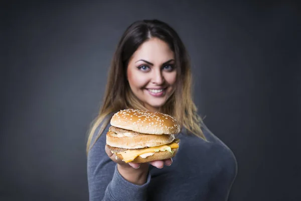Feliz hermosa joven modelo de tamaño grande posando con hamburguesa sobre un fondo de estudio gris, xxl mujer comiendo hamburguesa — Foto de Stock