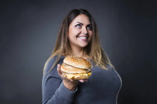 Feliz hermosa joven modelo de tamaño grande posando con hamburguesa sobre un fondo de estudio gris, xxl mujer comiendo hamburguesa — Foto de Stock