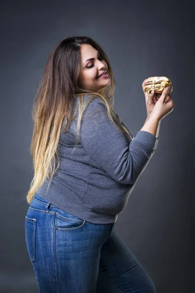 Feliz hermosa joven modelo de tamaño grande posando con hamburguesa sobre un fondo de estudio gris, xxl mujer comiendo hamburguesa — Foto de Stock