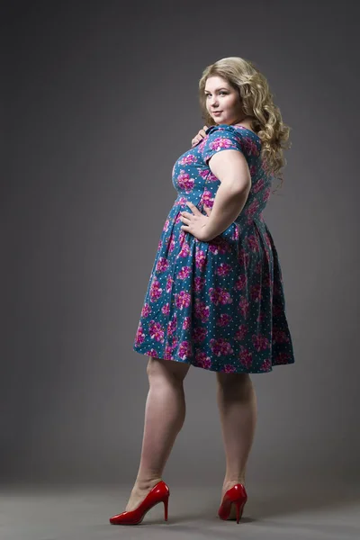 Young beautiful happy blonde plus size model in dress and shoes, xxl woman on gray studio background