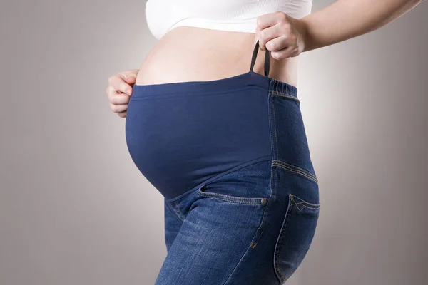 Беременная женщина в синих джинсах для беременных женщин на сером фоне — стоковое фото