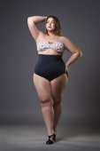 Plus-Size-Modell im sexy Badeanzug, junge dicke Frau auf grauem Hintergrund, übergewichtiger weiblicher Körper