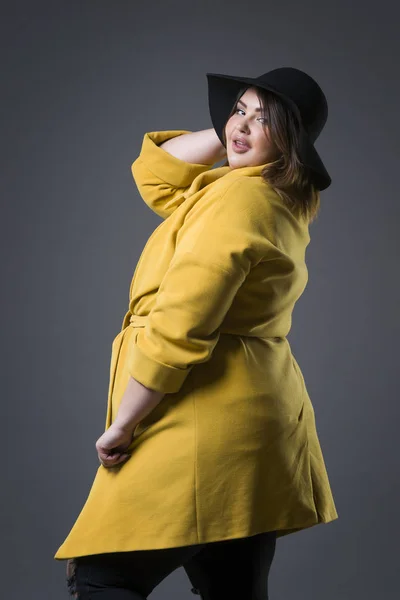Plus model de moda dimensiune în haină galbenă și pălărie neagră, femeie grasă pe fundal gri, corp feminin supraponderal — Fotografie, imagine de stoc