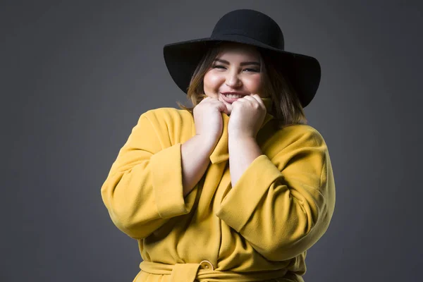 Plus size fashion model in yellow coat and black hat, fat woman on gray background, overweight female body — Stock Photo, Image