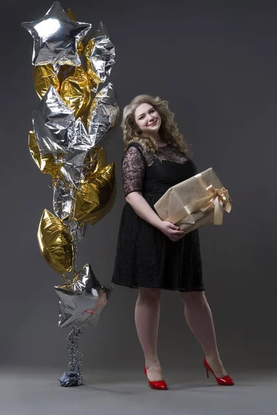 Plus femeie dimensiune în dres negru cu cutie cadou și baloane pe fundal gri — Fotografie, imagine de stoc