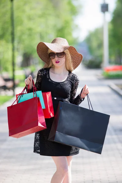 Snygg blond kvinna i svart klänning, solglasögon och hatt med shoppingkassar promenader i staden — Stockfoto