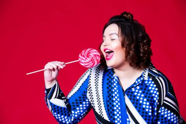 Mavi elbiseli, büyük lolipop yiyen, kırmızı arka planda şişman bir kadın.