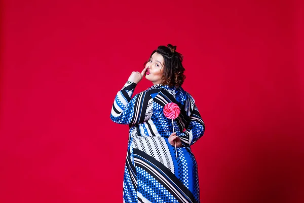 Модель plus size в синем платье с большим лоллипопом, толстая женщина на красном фоне — стоковое фото