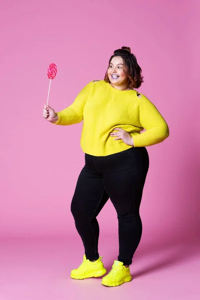 Alegre modelo de talla grande en ropa casual con piruleta grande, mujer gorda en jeans negros y jersey amarillo sobre fondo rosa — Foto de Stock