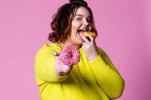 Alegre modelo más tamaño comer donas, mujer gorda come en exceso sobre fondo rosa — Foto de Stock