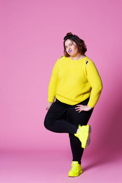 Cheerful plus size model in casual clothes, fat woman in black jeans and yellow jumper on pink background