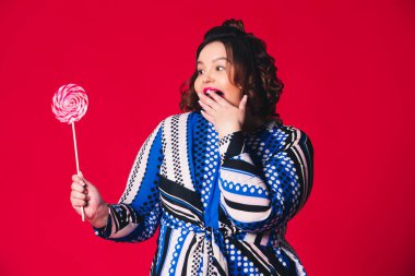 Artı geniş ağızlı, kırmızı arka planda büyük lolipop tutan bir model, vücut pozitifliği.