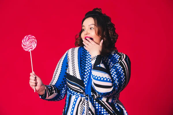 Artı geniş ağızlı, kırmızı arka planda büyük lolipop tutan bir model, vücut pozitifliği.