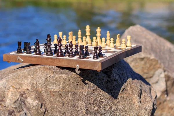 Chess board with chess pieces on rock with river embankment back — Stock Photo, Image