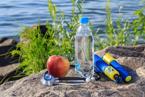 Jump rope, apple, stethoscope and bottle with water on rock with — Stock Photo, Image